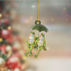 May include: A Christmas ornament featuring two green frogs holding a green umbrella. The frogs are adorned with colorful string lights and the text "Name 1" and "Name 2". The ornament hangs from a gold string.
