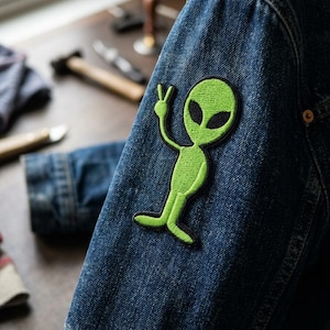 May include: A green alien patch with black eyes and a peace sign hand gesture is sewn onto a blue denim jacket sleeve. The alien is outlined in black. The jacket is made of blue denim.