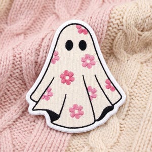 May include: An embroidered patch shaped like a ghost with a cream-colored base and a pattern of small pink flowers. The ghost features two solid black circular eyes and is outlined with a thick white border and thin black stitching details.