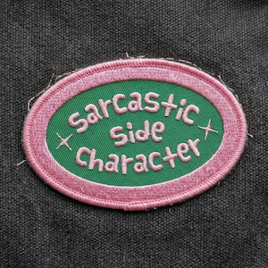 May include: An embroidered patch with the words "Sarcastic Side Character" in white, inside a green oval with a pink border. Two pink crosses are on either side of the text. The patch is on a dark gray fabric background.