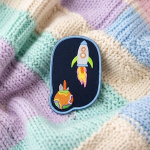 May include: A dark blue embroidered patch with a light blue border featuring a white rocket ship with green fins and a yellow and orange flame, positioned above a round orange planet with green and purple surface details and a small gray antenna.