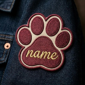May include: Embroidered paw print patch in burgundy, cream, and gold, with the word "name" in a cursive font. The patch is sewn onto a dark blue denim jacket, showcasing a personalized pet accessory.