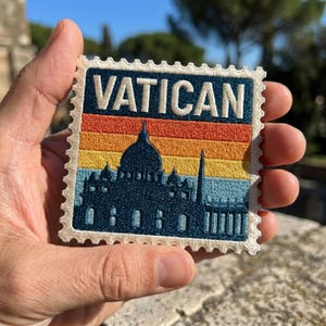 May include: A square embroidered patch shaped like a postage stamp with a scalloped edge. It features the word VATICAN in white letters above a silhouette of St. Peter's Basilica against a horizontal gradient background of orange, yellow, and blue.