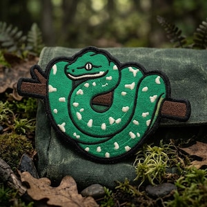 May include: Embroidered patch featuring a green snake with white spots coiled around a brown branch. The snake has a black outline and is set against a backdrop of green foliage and moss.