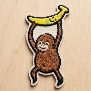 May include: Embroidered patch featuring a brown monkey holding a yellow banana overhead. The monkey has a smiling face and is outlined in black. The banana has a green tip and a smiling face. The patch is on a beige background.