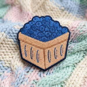May include: An embroidered patch featuring a square tan basket filled with a cluster of dark blue blueberries. The design is outlined in a dark navy blue thread, showcasing a detailed stitch pattern that defines the basket and the fruit.