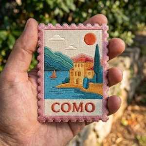 May include: A rectangular embroidered patch shaped like a postage stamp with a pink scalloped border. The design features a yellow building by a blue lake, green mountains, a tall cypress tree, an orange sun, and the word COMO in orange letters.