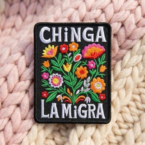 May include: A rectangular black embroidered patch featuring a colorful floral arrangement with green stems and leaves. The white text CHINGA is embroidered at the top, and the white text LA MIGRA is embroidered at the bottom of the patch.