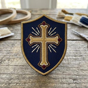 May include: Embroidered shield-shaped patch with a gold cross design on a navy blue background. The cross is detailed with white rays and burgundy accents. Embroidery floss, scissors, and wooden hoops are in the background.