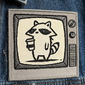May include: Embroidered patch featuring a cartoon raccoon holding a coffee cup, set within a retro television frame. The patch is in black, gray, and white, with a denim background. The design includes a volume dial and a channel dial.