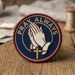 May include: Round embroidered patch with the words "PRAY ALWAYS" encircling a design of praying hands and a gold cross. The patch has a navy blue background with gold and burgundy accents.