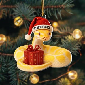 May include: A yellow snake ornament wearing a red Santa hat with the name "Cherry" on it. The snake is coiled around a red gift box with a red bow and gold stars. The background is a blurred Christmas tree.