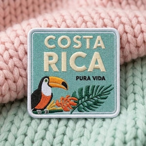 May include: A square embroidered patch with a light blue background featuring the text COSTA RICA in large cream letters and PURA VIDA in smaller black letters. A toucan, orange flowers, and a green palm leaf are embroidered at the bottom.