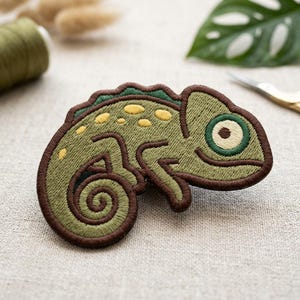 May include: Embroidered chameleon patch in shades of green and brown. The patch features a detailed design with yellow spots and a curled tail. The chameleon has a large, round eye. Perfect for adding to clothing or accessories.