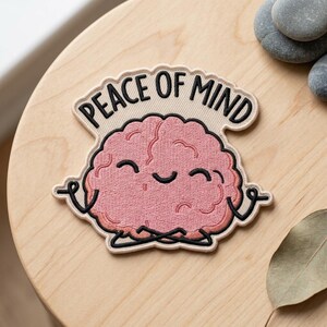 May include: Embroidered patch featuring a pink brain in a meditative pose. The patch has the words "PEACE OF MIND" arched above the brain. The brain has a smiling face and is outlined in black. The patch is on a light beige background.