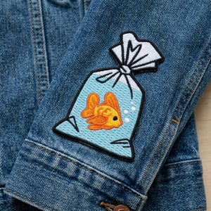 May include: Close-up of a denim jacket sleeve with an embroidered patch. The patch features a goldfish in a plastic bag with a light blue background and white accents. The goldfish is orange and yellow.