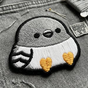 May include: Embroidered patch featuring a cartoon bird with gray and white feathers, black eyes, and yellow heart-shaped feet. The patch is outlined in black and sits on gray denim. A small "KIDS TV" label is visible.