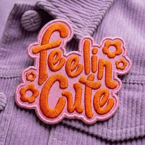 May include: A pink and orange embroidered patch with the words "Feelin' Cute" in a playful font. The patch is surrounded by small orange flower designs and is set against a purple corduroy fabric.