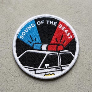 May include: Embroidered patch with a police car design. The patch features the text "SOUND OF THE BEAST" in a circular pattern around the image of a police car with flashing red and blue lights. The patch is black, white, red, and blue.