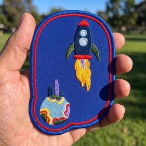 May include: A blue embroidered patch with a red border featuring a dark blue rocket ship with green fins and a yellow flame trail, positioned above a multicolored planet with purple, yellow, green, and red swirling patterns on a light blue base.