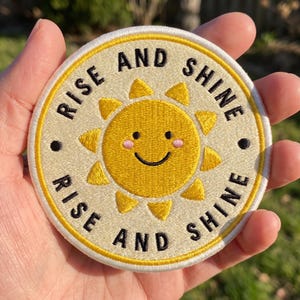 May include: A circular embroidered patch featuring a yellow sun with a smiling face and pink cheeks at the center. The background is cream-colored with a yellow border. The text "RISE AND SHINE" is stitched in black around the sun in a circular pattern.
