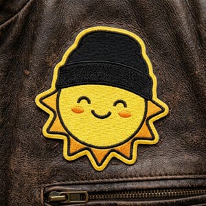 May include: Embroidered patch featuring a smiling sun wearing a black beanie. The sun is yellow with orange rays and rosy cheeks. The patch is sewn onto a brown leather jacket.