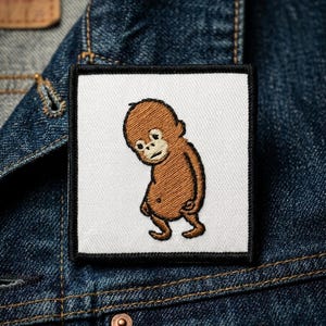 May include: Embroidered patch featuring a cartoon orangutan in shades of brown, with a white background and a black border. The patch is square-shaped and is set against a denim jacket.