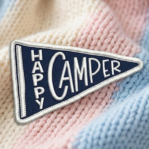 May include: A triangular navy blue felt pennant featuring white embroidered text that reads HAPPY CAMPER. The pennant has a white stitched border and is positioned against a background of soft, chunky knit fabric in pastel stripes.