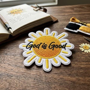 May include: A white and yellow embroidered patch with the text "God is Good" in black script. The patch is in the shape of a sun. Also pictured are a journal, pen, thread, scissors, and a dried flower on a wooden surface.