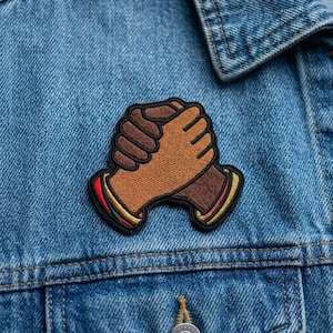 May include: Embroidered patch featuring two hands clasped together in a gesture of unity. The patch has brown and tan skin tones, with red, yellow, and black accents. The patch is sewn onto a blue denim jacket.