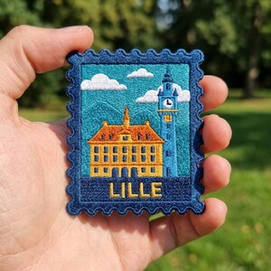 May include: A rectangular embroidered patch shaped like a postage stamp with a dark blue scalloped border. The design features a yellow building with an orange roof and a tall blue clock tower against a teal sky with white clouds. The word LILLE is stitched in yellow at the bottom.