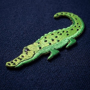 May include: A green embroidered alligator patch with black spots and details. The patch is on a navy blue textured background. The alligator is facing left, with its tail curved. The patch is likely for clothing or accessories.