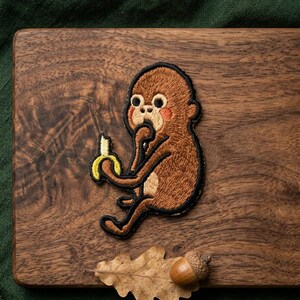 May include: Embroidered patch featuring a cartoon monkey holding a banana. The monkey is brown with a black outline and red cheeks. The patch is on a wooden surface with an acorn and leaf.