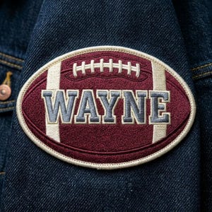 May include: An embroidered patch in the shape of a football, with the name "WAYNE" in gray letters. The football is maroon with white stripes and stitching, and is set against a dark blue denim jacket.
