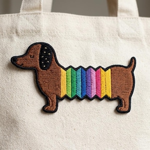 May include: Embroidered patch of a dachshund dog with a rainbow-colored accordion-style body. The dog is brown with a black outline and black accents on its head. The patch is on a beige canvas tote bag.