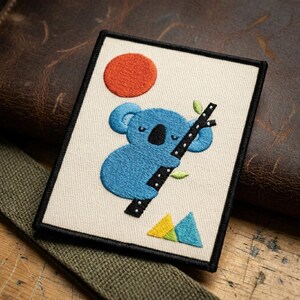 May include: Embroidered patch featuring a blue koala clinging to a branch, with an orange sun and geometric shapes. The patch has a black border and is set against a neutral background.