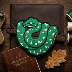 K&ouml;nnte beinhalten: Ein gr&uuml;ner Schlangenaufn&auml;her mit wei&szlig;en Punkten auf einem braunen Ast liegt auf einem Lederjournal. Eine Lupe, Garnrollen, ein kleines Buch mit "OBSERVATIONS: SERPENTES" und ein getrocknetes Blatt sind ebenfalls vorhanden.