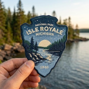 May include: An embroidered patch shaped like an arrowhead featuring a landscape of Isle Royale National Park in Michigan. The design includes a lake, pine trees, a large moon, and stars in shades of blue, white, and cream with the text 1940.
