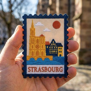 May include: A rectangular embroidered patch shaped like a postage stamp with a dark blue border. It features a yellow cathedral, blue half-timbered houses, a red sun, and white clouds against a cream background. The text STRASBOURG is in red.