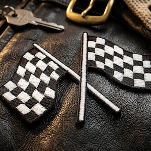 May include: Two embroidered checkered flag patches with black and white squares, set against a textured black leather surface. A key, a gold buckle, and a tan glove are also visible.