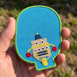May include: A bright blue, oval-shaped embroidered patch featuring a tan robot character with a dark blue antenna, two red claw hands, and a yellow base. The patch is outlined with a vibrant lime green border and has a smooth fabric texture.