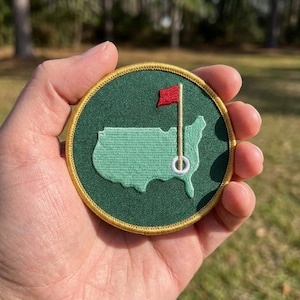 May include: A circular embroidered patch featuring a dark green background with a light green map of the United States. A white golf hole with a thin tan pole and a red flag is positioned over the map. The patch has a gold stitched border.