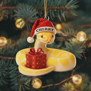 May include: A yellow snake ornament wearing a red Santa hat with the name "Cherry" on it. The snake is holding a red gift box with a red bow and star details. The ornament is hanging in front of a Christmas tree.
