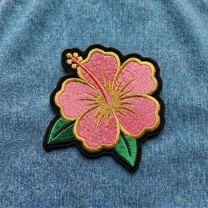 May include: Embroidered pink hibiscus flower patch with yellow and green accents, and a black border. The patch is on a blue denim background. The flower has a long pink stamen and green leaves.
