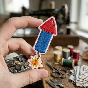 May include: An embroidered patch shaped like a firework rocket with a red triangular top, a blue rectangular body, and a yellow and orange starburst at the bottom, all outlined in white thread with a brown fuse connecting the body to the starburst.