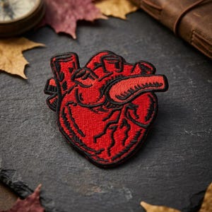 May include: A red embroidered patch in the shape of a human heart with black outlining. The patch is on a dark gray surface, surrounded by autumn leaves, a compass, and a leather-bound journal.