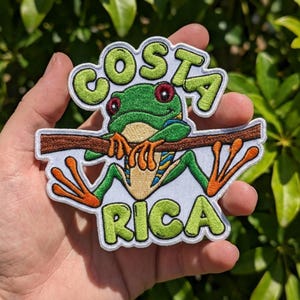 May include: An embroidered patch featuring a green red-eyed tree frog with orange feet clinging to a brown branch. The text COSTA RICA is stitched in lime green letters above and below the frog against a white background with a white border.