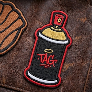May include: Embroidered patch featuring a black spray paint can design with a red outline and the word "TAG" in red. The top of the can is red, and the nozzle is gold. The patch is on a brown leather surface.