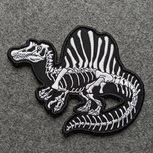May include: Black and white embroidered patch featuring a Spinosaurus dinosaur skeleton. The patch has a detailed design of the dinosaur's bones on a black background, with a white outline. The patch is likely for clothing or accessories.