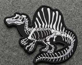 May include: Black and white embroidered patch featuring a Spinosaurus dinosaur skeleton. The patch has a detailed design of the dinosaur's bones on a black background, with a white outline. The patch is likely for clothing or accessories.